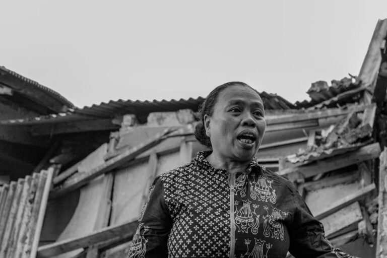 Black and white portrait of a woman standing in front of a damaged house, Cianjur, Indonesia.