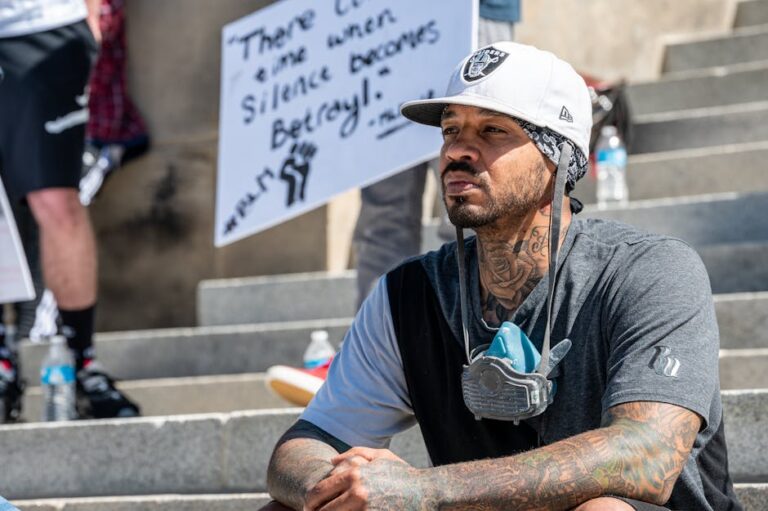 A thoughtful man sits on stairs during a peaceful protest, wearing a mask and displaying tattoos.