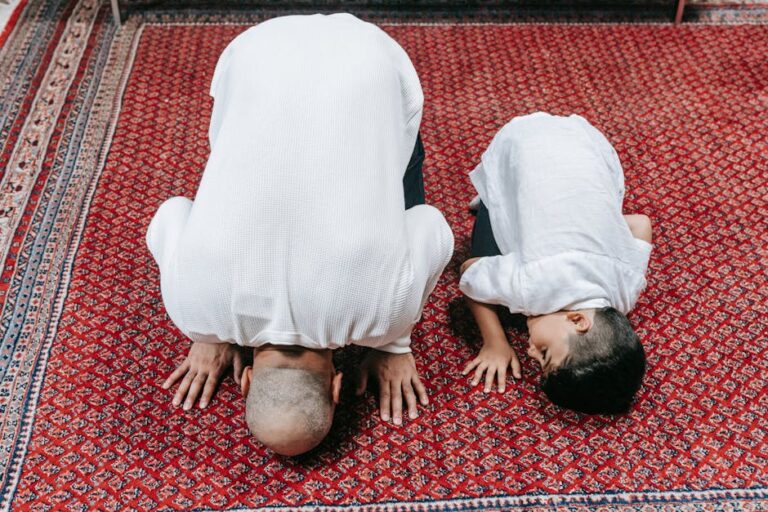 A father and son kneeling in prayer on a beautifully patterned red carpet, symbolizing religious devotion.