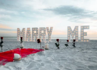 Ideias criativas para pedido de casamento na praia Beach proposal setup with roses and illuminated 'Marry Me' sign at sunset.