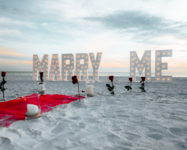 Beach proposal setup with roses and illuminated 'Marry Me' sign at sunset.