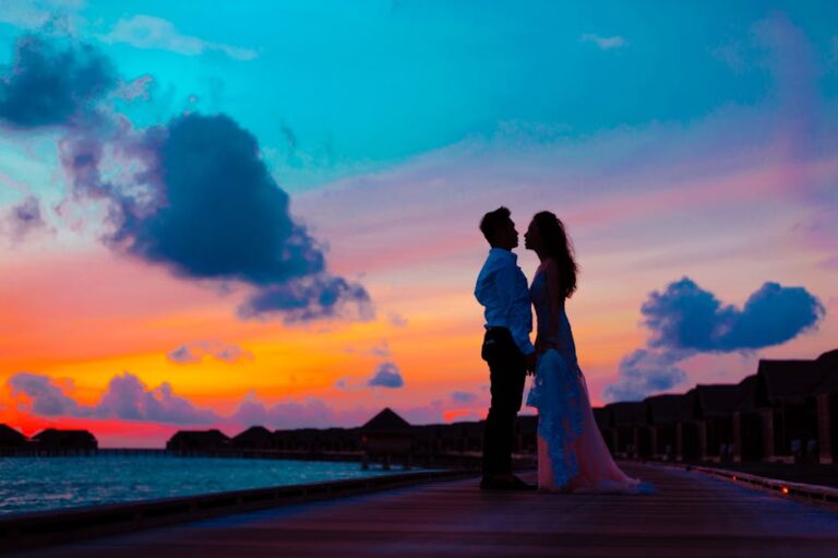 Silhouette of a couple embracing against a vibrant tropical sunset on a beach pier.
